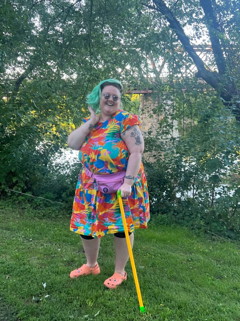 Jesse stands jauntily outdoors wearing a bright printed poplin dress, knee braces, and a pink fanny pack. Their hair is blue and she's wearing sunglasses and standing with a marigold cane with neon lime handles and ferrule.