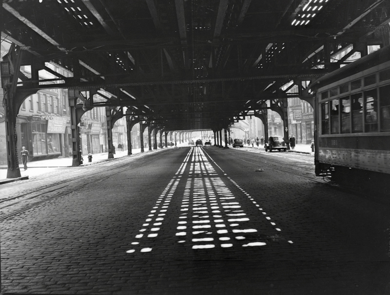 A black and white photo by Weegee of the elevated subway lines with a streetcar passing underneath.