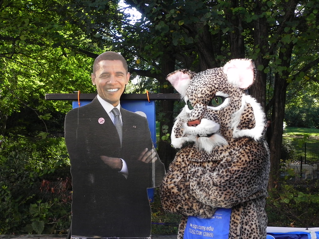 A photo of Lexington Lynx, CUNY SPS' mascot, posing in a park with a cardboard cutout of Barack Obama smiling with his arms crossed.