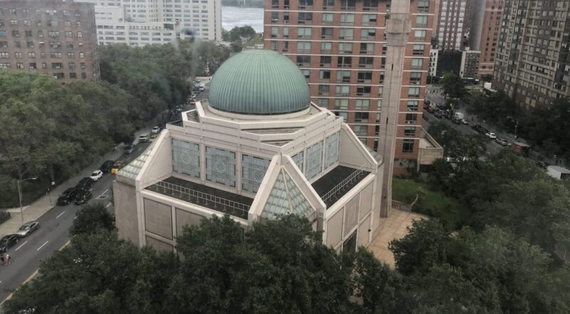 Mint dome that sits on top of a building. this is a famous mosque located in manhattan. lush trees sit at the bottom and behind the mosque you can see city traffic and apartment buildings.