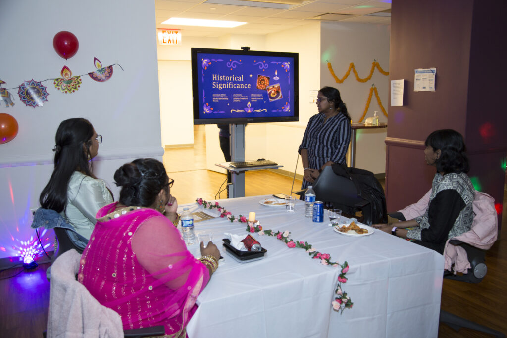3 people are sitting around a rectangle table. on person is standing at the head off the table giving a presentation about Diwali